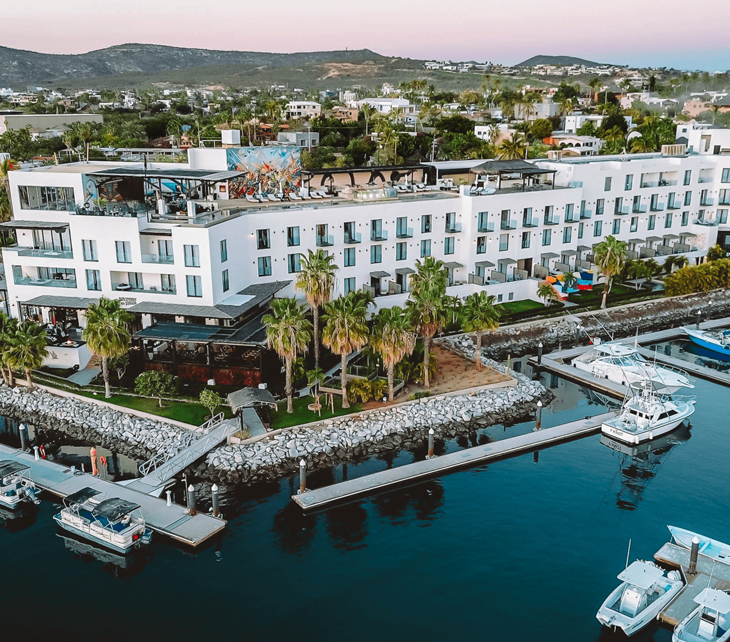 A modern white waterfront hotel with balconies, surrounded by palm trees and docks with boats in a marina.