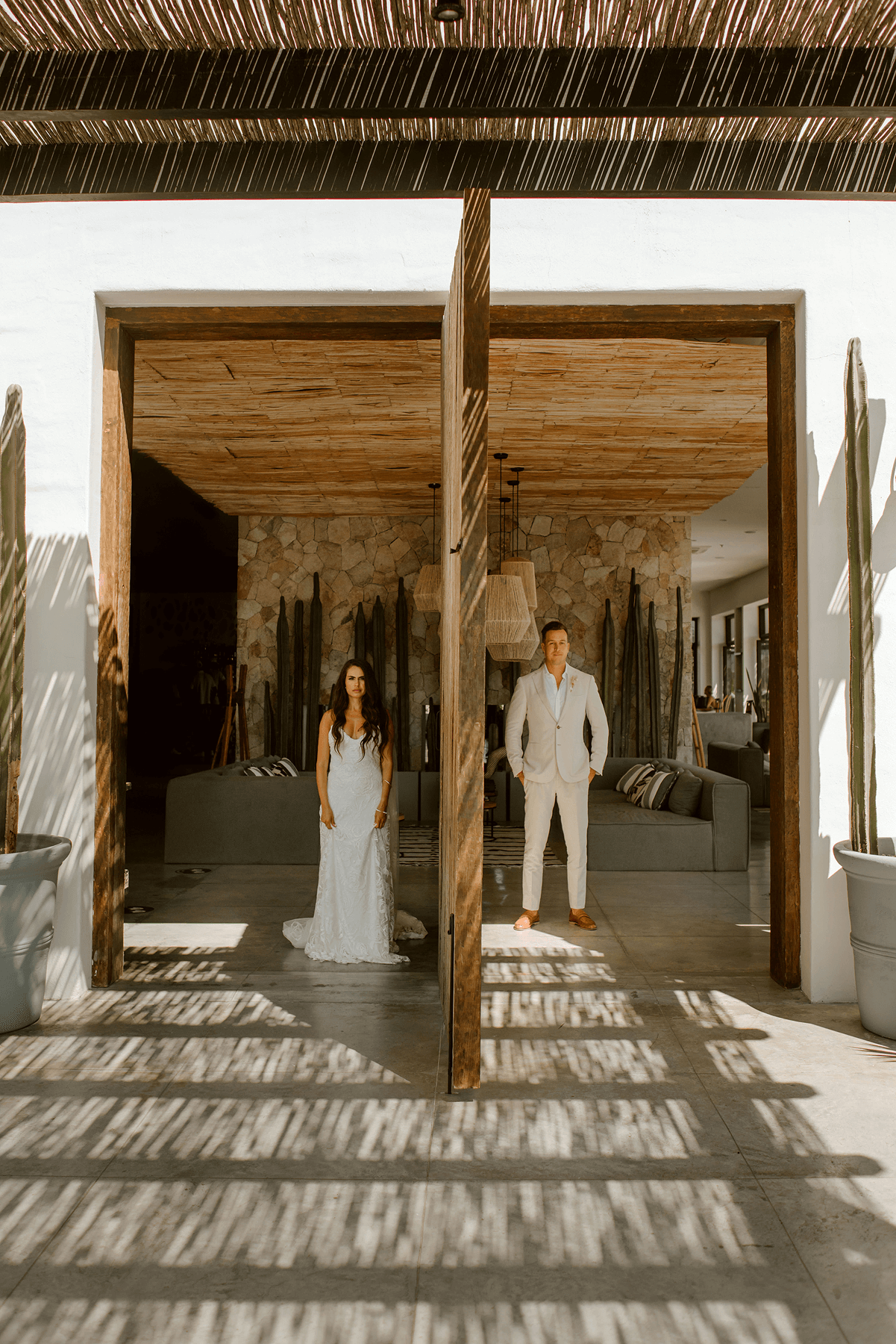 Bride in a white wedding dress and groom in a light suit standing separately inside a modern building, framed by wooden doors with patterned shadows on the floor.