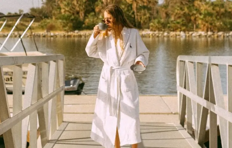 Woman in white bathrobe drinking from a cup on a dock by a calm lake in the morning.