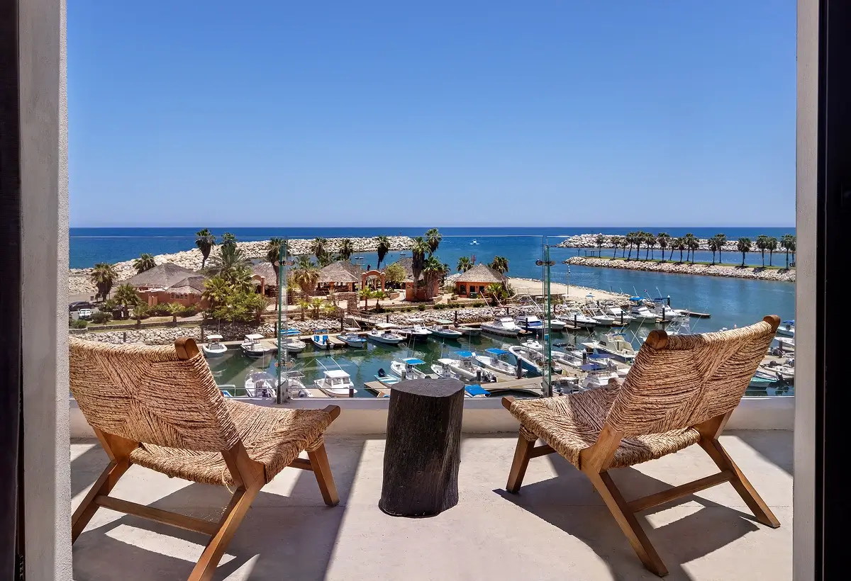 View from a balcony with two woven chairs and a small table overlooking a marina with boats, palm trees, and the ocean under a clear blue sky.