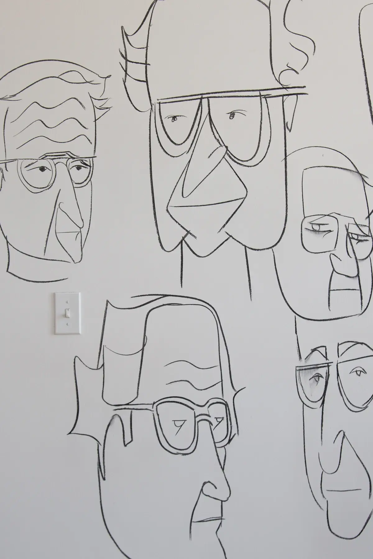 Black line drawings of abstract human faces with glasses on a white wall, with a light switch visible on the left.