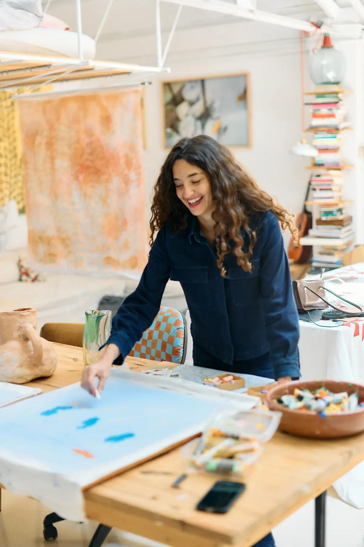 Smiling woman with curly hair painting blue shapes on a canvas in a bright, art-filled workspace.