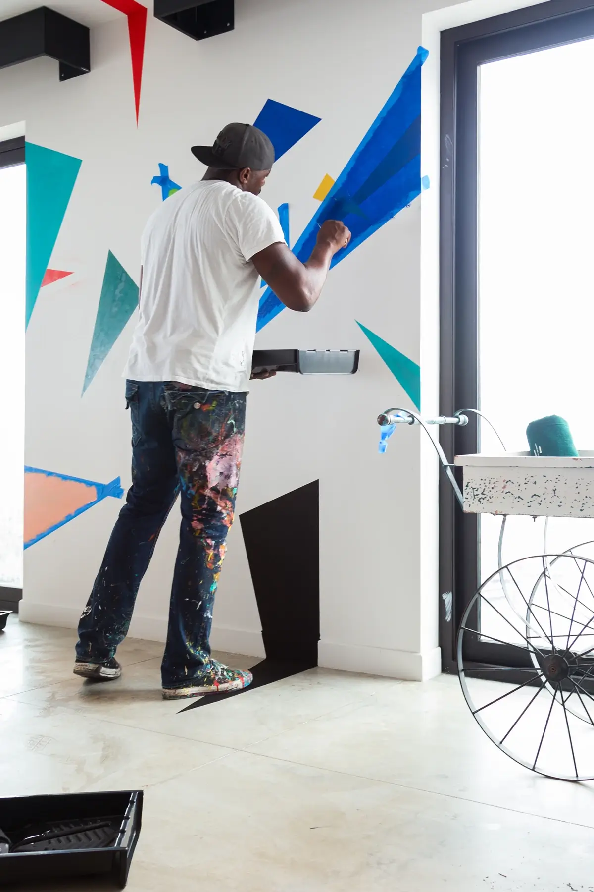 Male artist wearing a backwards cap and paint-splattered pants painting blue geometric shapes on a white wall near a window.