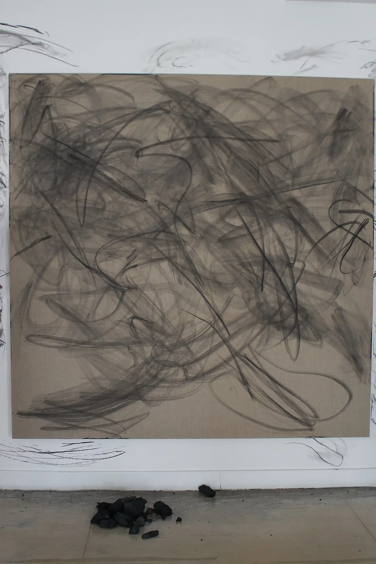Abstract black charcoal scribbles on a large beige canvas mounted on a white wall with charcoal pieces scattered on the floor beneath.