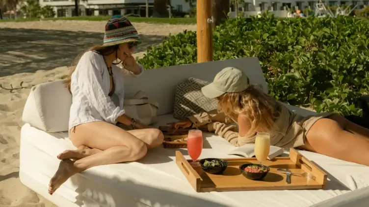 Two women relaxing on a cushioned outdoor daybed at the beach, playing a board game with drinks and snacks on a wooden tray nearby.