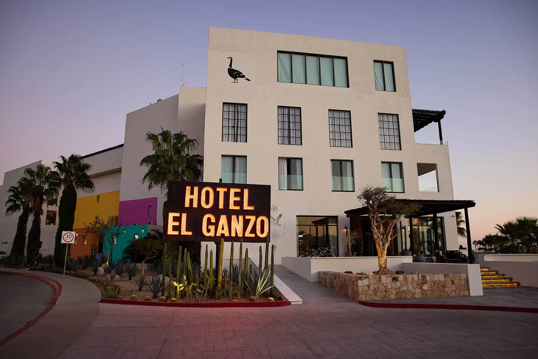 Modern white hotel building with large illuminated sign reading 'Hotel El Ganzo' at dusk.