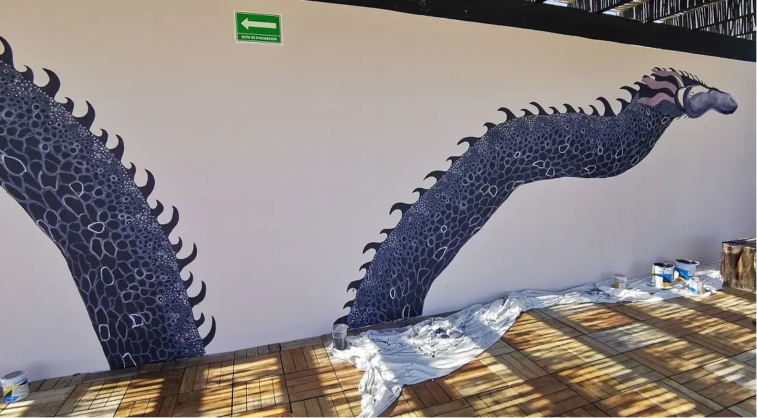 Wall mural of a large, segmented sea creature or serpent with dark scales and curved fins, partially painted on a white wall above wooden floor with painting supplies nearby.