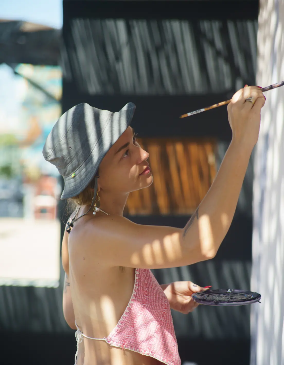 Woman wearing a gray bucket hat and pink top painting on a vertical surface while holding a paint plate.
