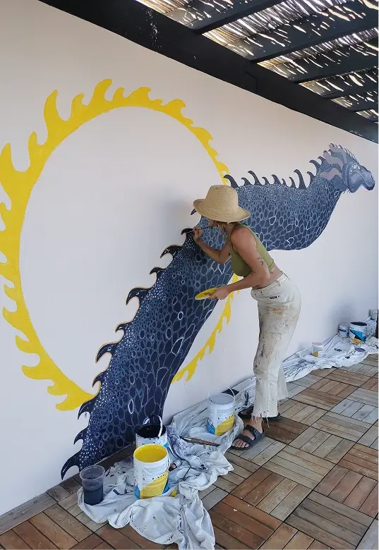 Woman wearing a hat painting a large dark blue dragon-like creature with a yellow flame circle on a white wall.