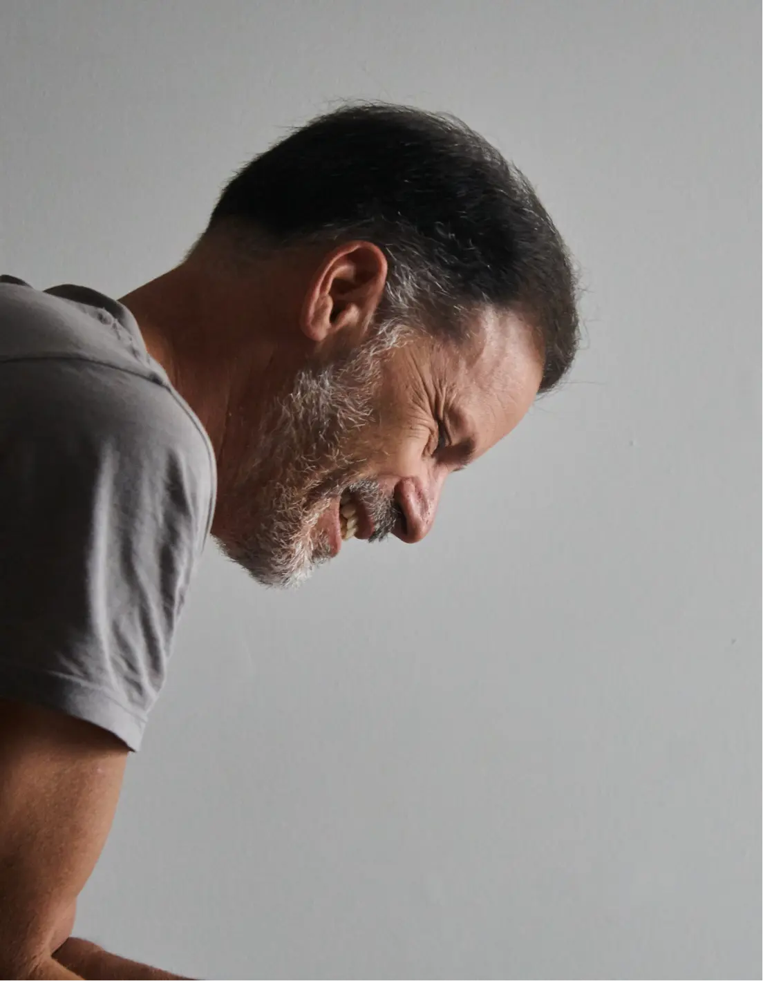 Perfil lateral de un hombre de mediana edad con cabello oscuro y barba gris sonriendo contra un fondo gris liso.