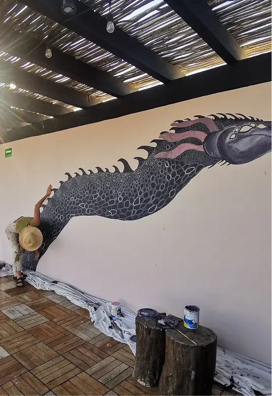 Person with a large straw hat painting a detailed mural of a black and purple sea creature on a white wall under a wooden pergola.