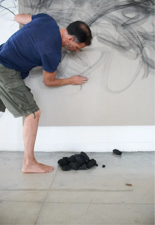 Hombre con camisa azul y pantalones cortos verdes dibujando en la pared con carboncillo mientras está descalzo.