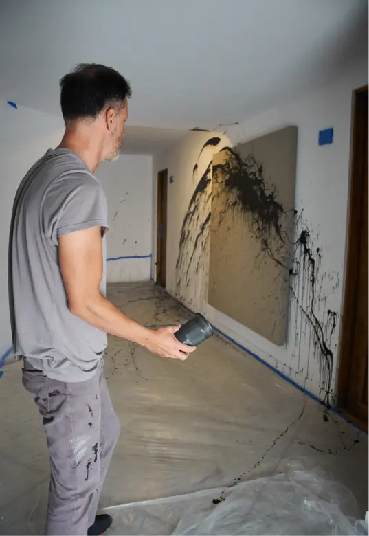 Un hombre vestido de gris utiliza un secador de pelo para esparcir salpicaduras de pintura negra sobre un gran lienzo colgado en la pared de un pasillo.