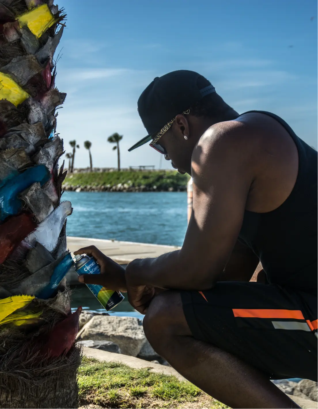Hombre con gafas de sol y gorra negra pintando con spray el tronco de una palmera cerca de la costa.