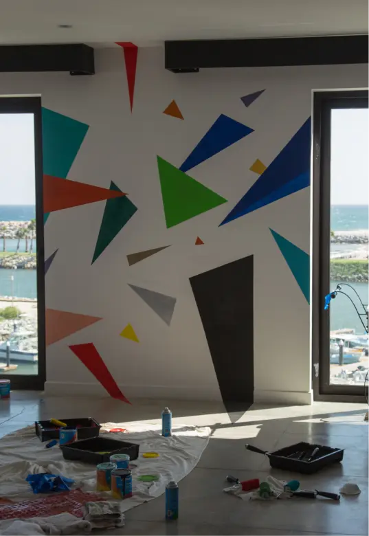 Pared con formas geométricas pintadas de colores entre dos grandes ventanas con vistas a un puerto deportivo, con materiales de pintura en el suelo.