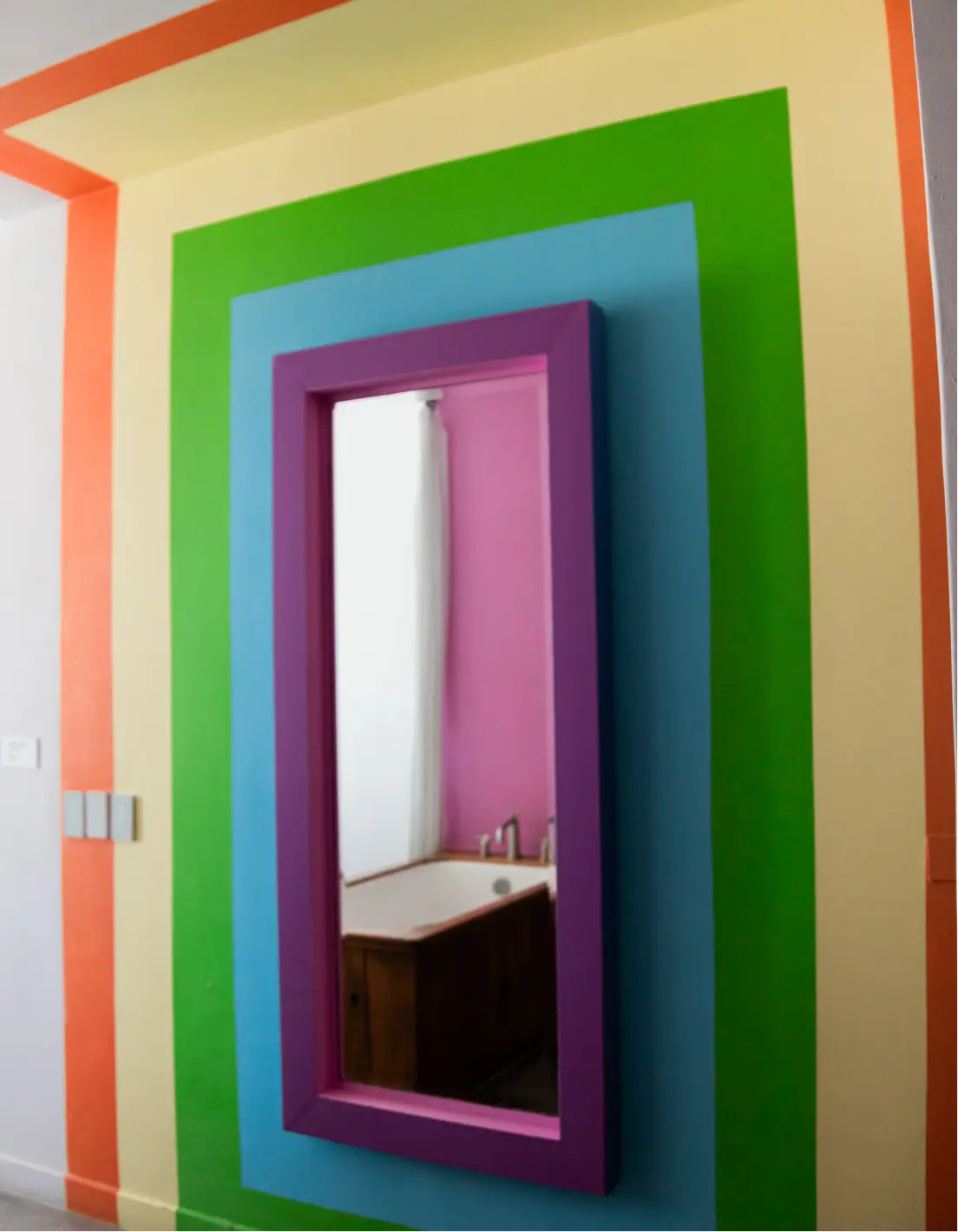 Wall painted with vertical stripes in orange, yellow, green, and blue surrounding a purple-framed mirror reflecting a bathtub and pink wall.