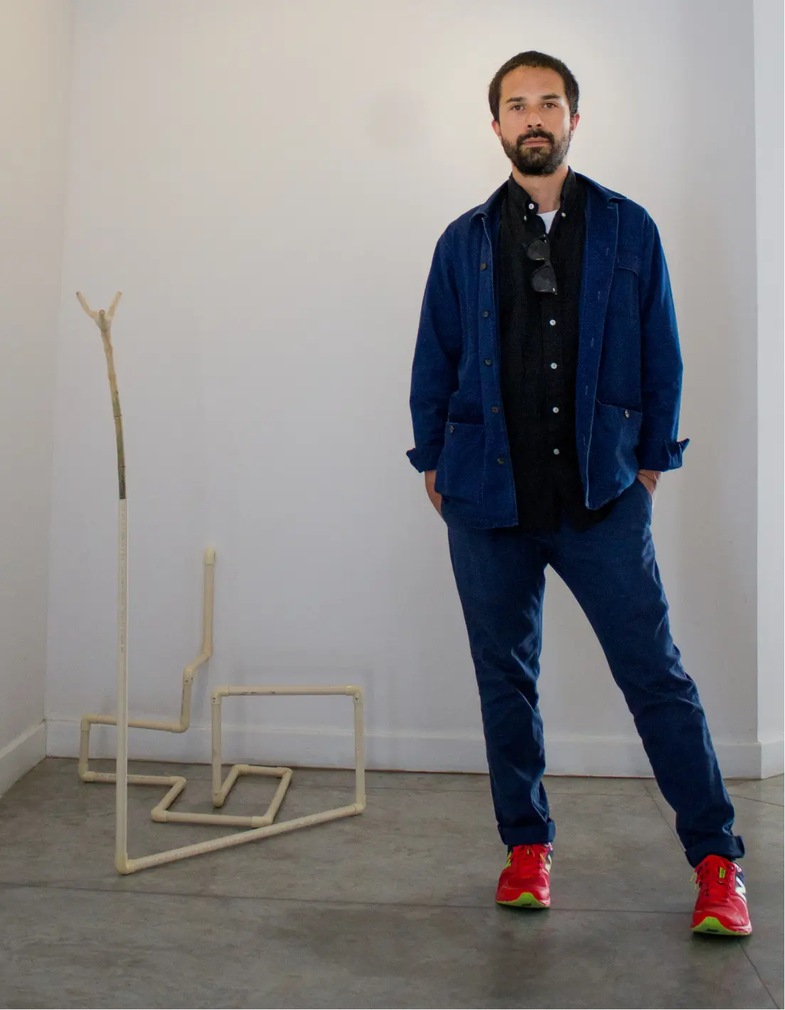 Man with beard wearing a blue jacket, black shirt, blue pants, and red sneakers standing in an art gallery next to a minimalist sculpture made of beige pipes.