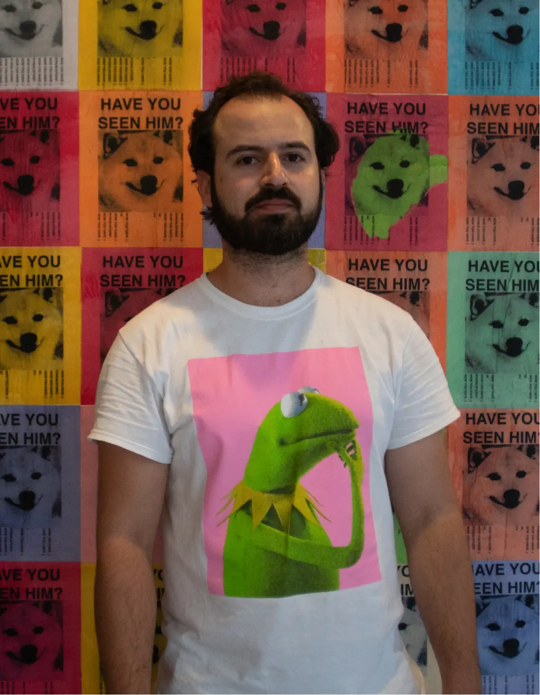Man with beard wearing a white T-shirt featuring Kermit the Frog against a colorful wall of 'Have You Seen Him?' dog posters.