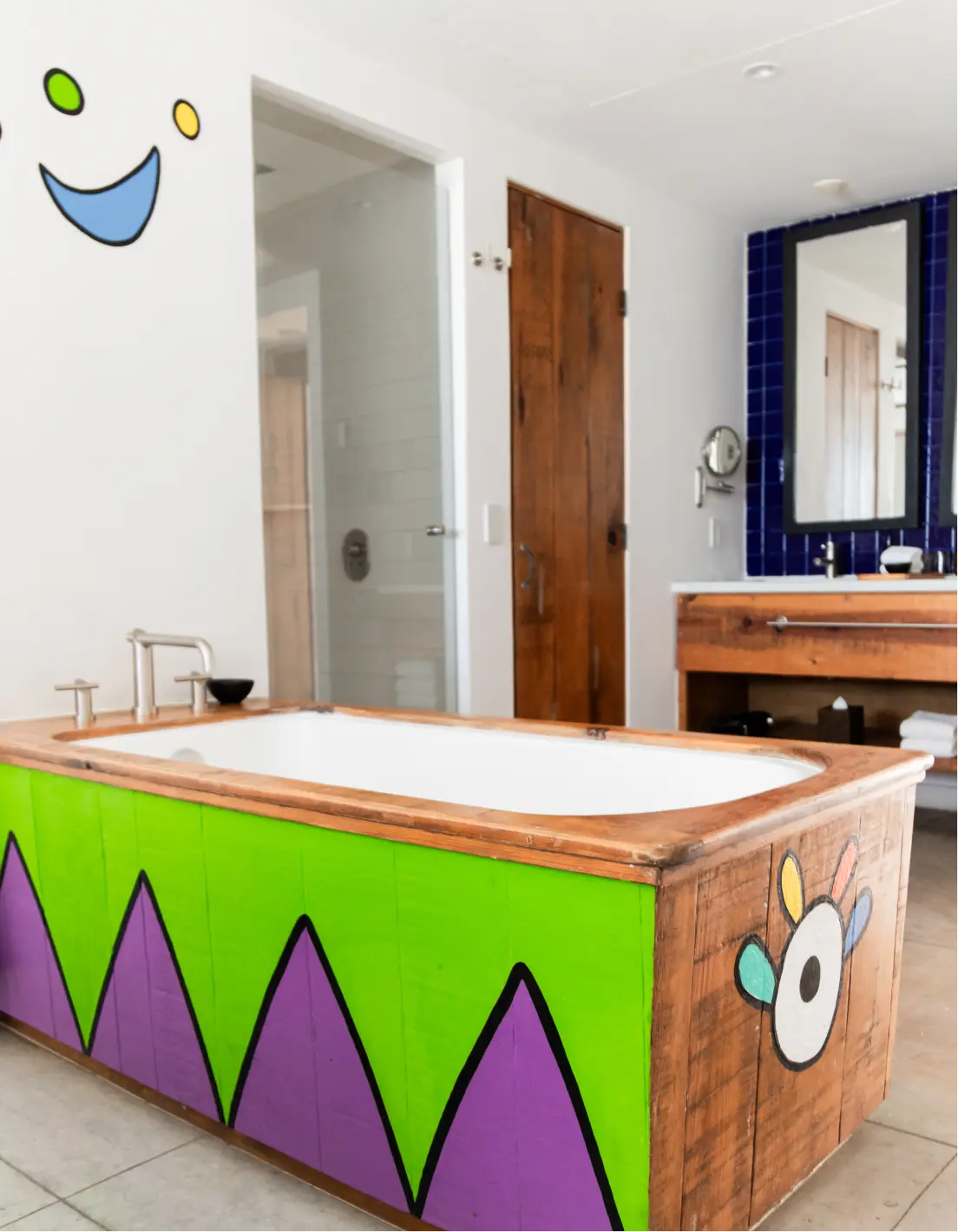 Bathroom with a wooden bathtub featuring bright green and purple geometric patterns and colorful painted accents, next to a wooden door and a vanity with a mirror on a blue tiled wall.