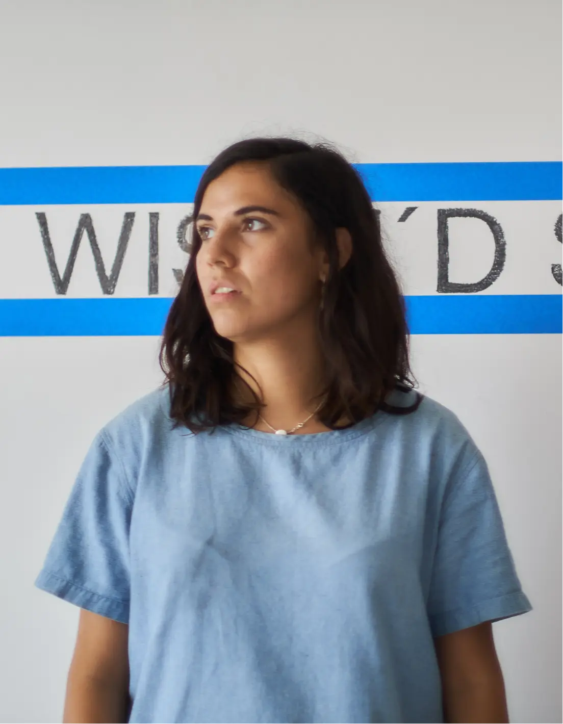 Woman with dark hair wearing a light blue shirt looking to the side against a white wall with blue stripes and partially visible large text.
