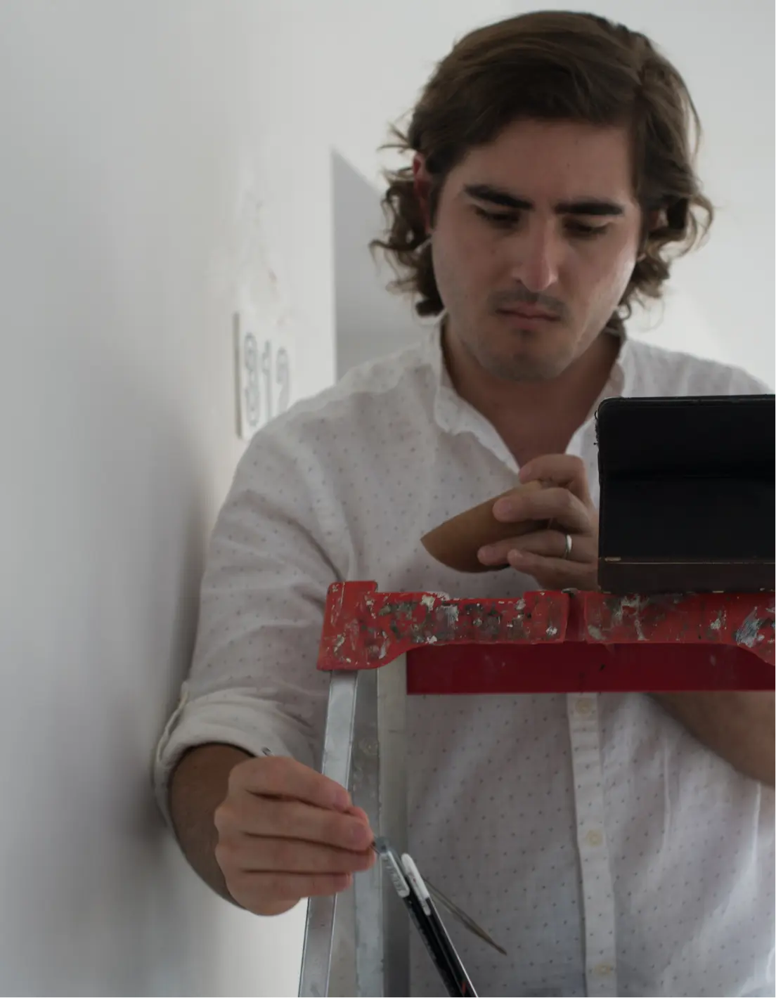 Man in a white shirt working on a ladder, holding a paintbrush and a sanding block.