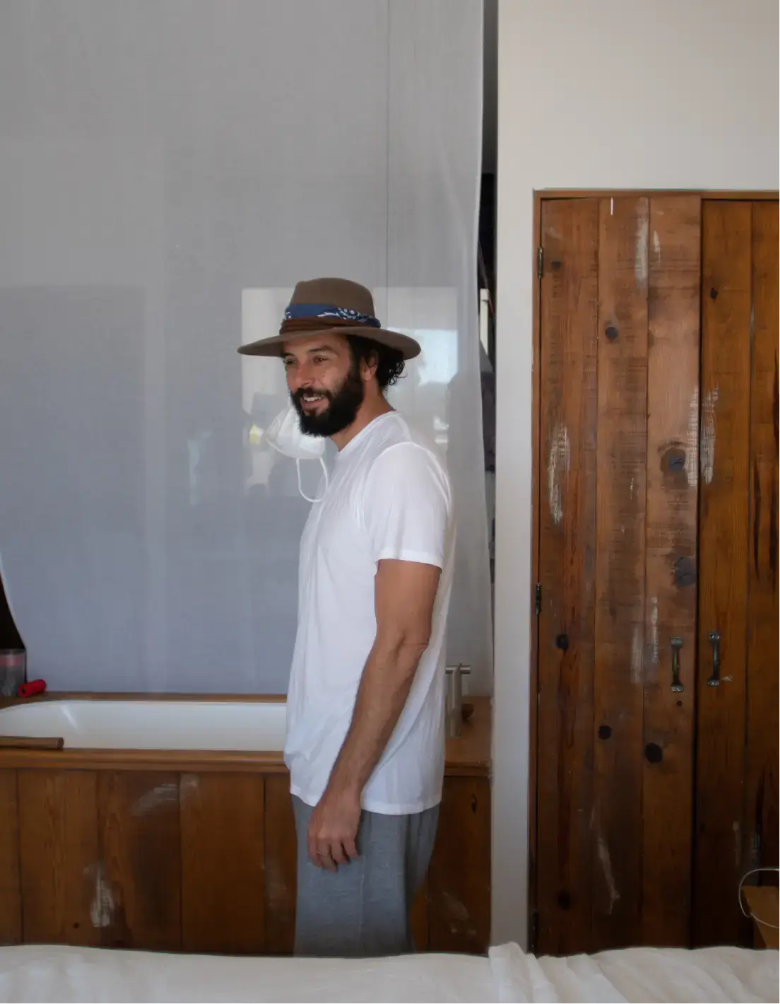 Hombre con barba, sombrero marrón y camiseta blanca, de pie de lado en una habitación con armarios de madera y una bañera.