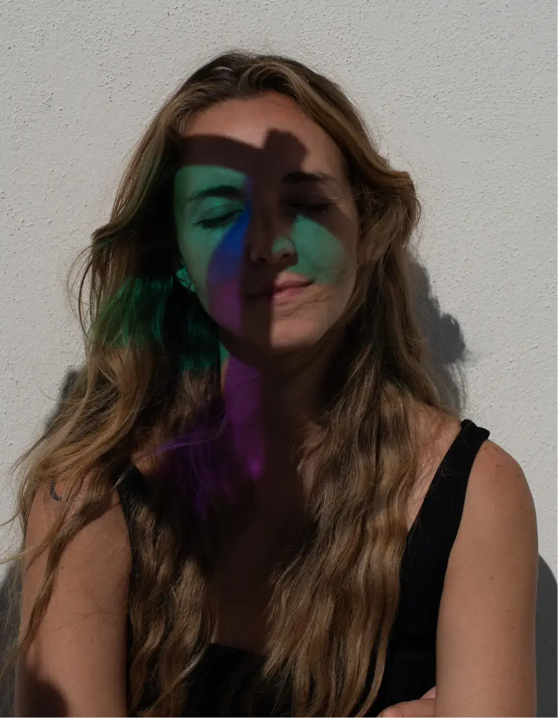 Mujer con cabello largo y ondulado y ojos cerrados, con sombras coloridas proyectadas en su rostro contra una pared blanca con textura.