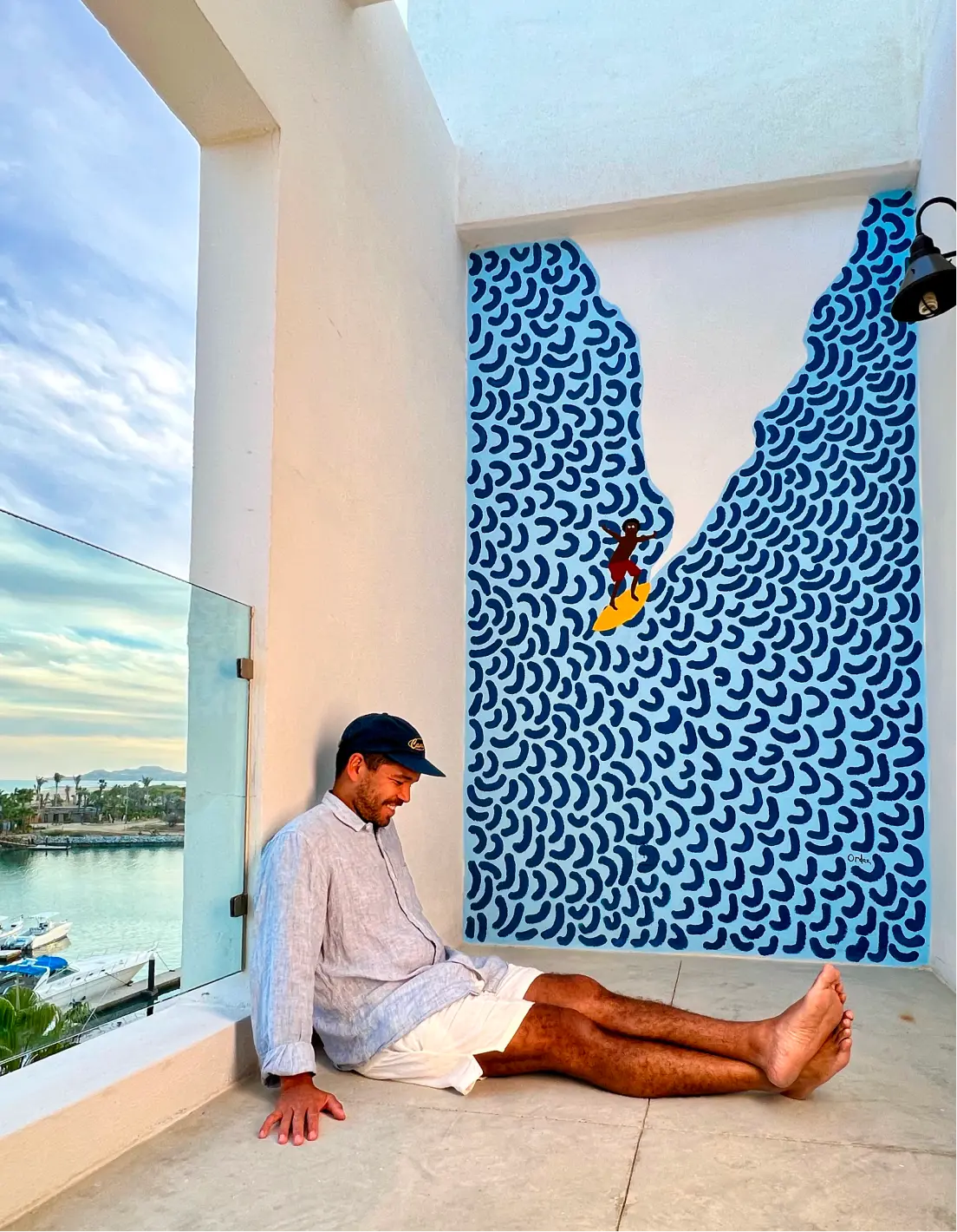 Man in light shirt and white shorts sitting on floor near wall with blue abstract mural of surfer riding a yellow surfboard.