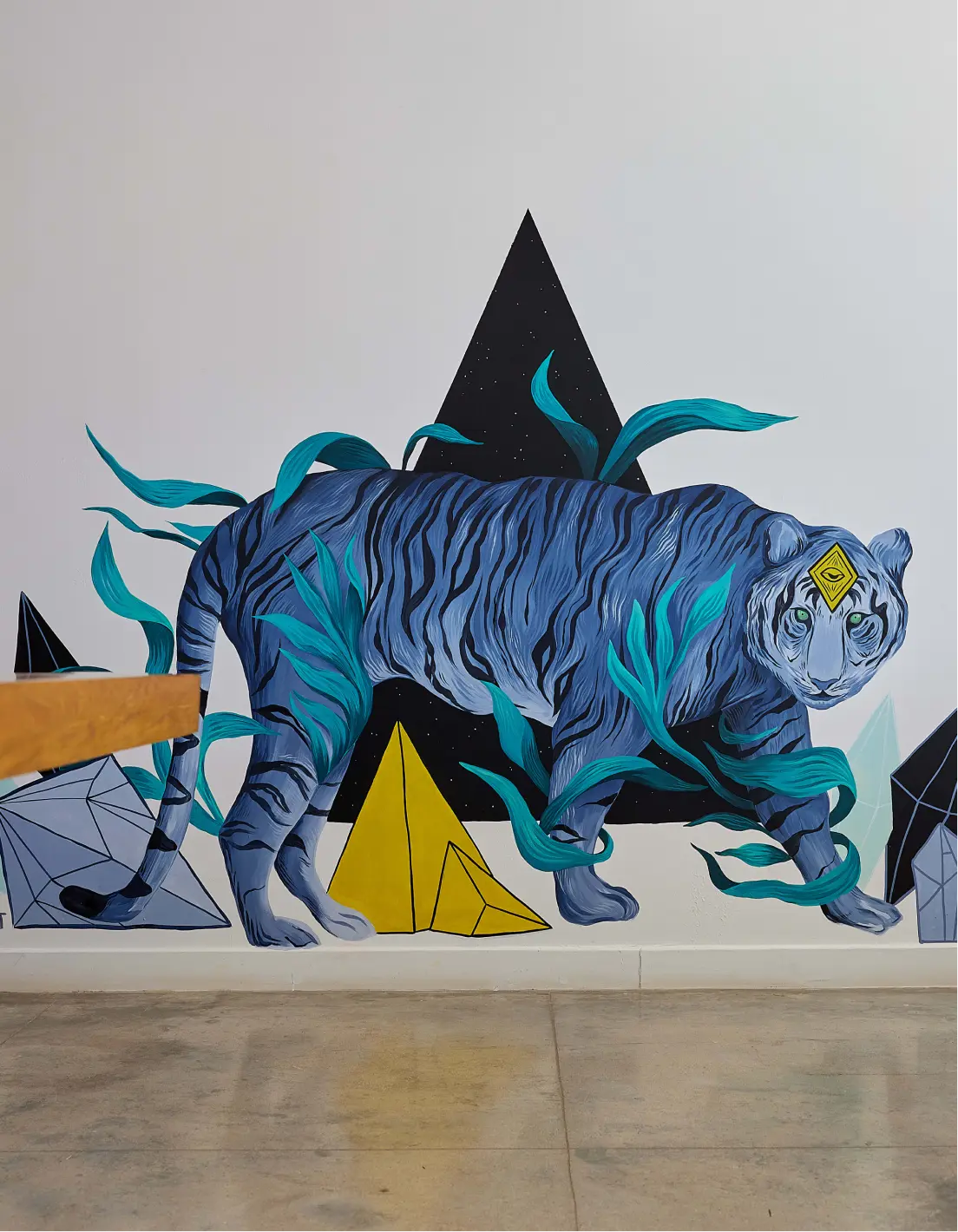 Wall mural of a blue tiger surrounded by teal leaves and geometric shapes including black and yellow pyramids.