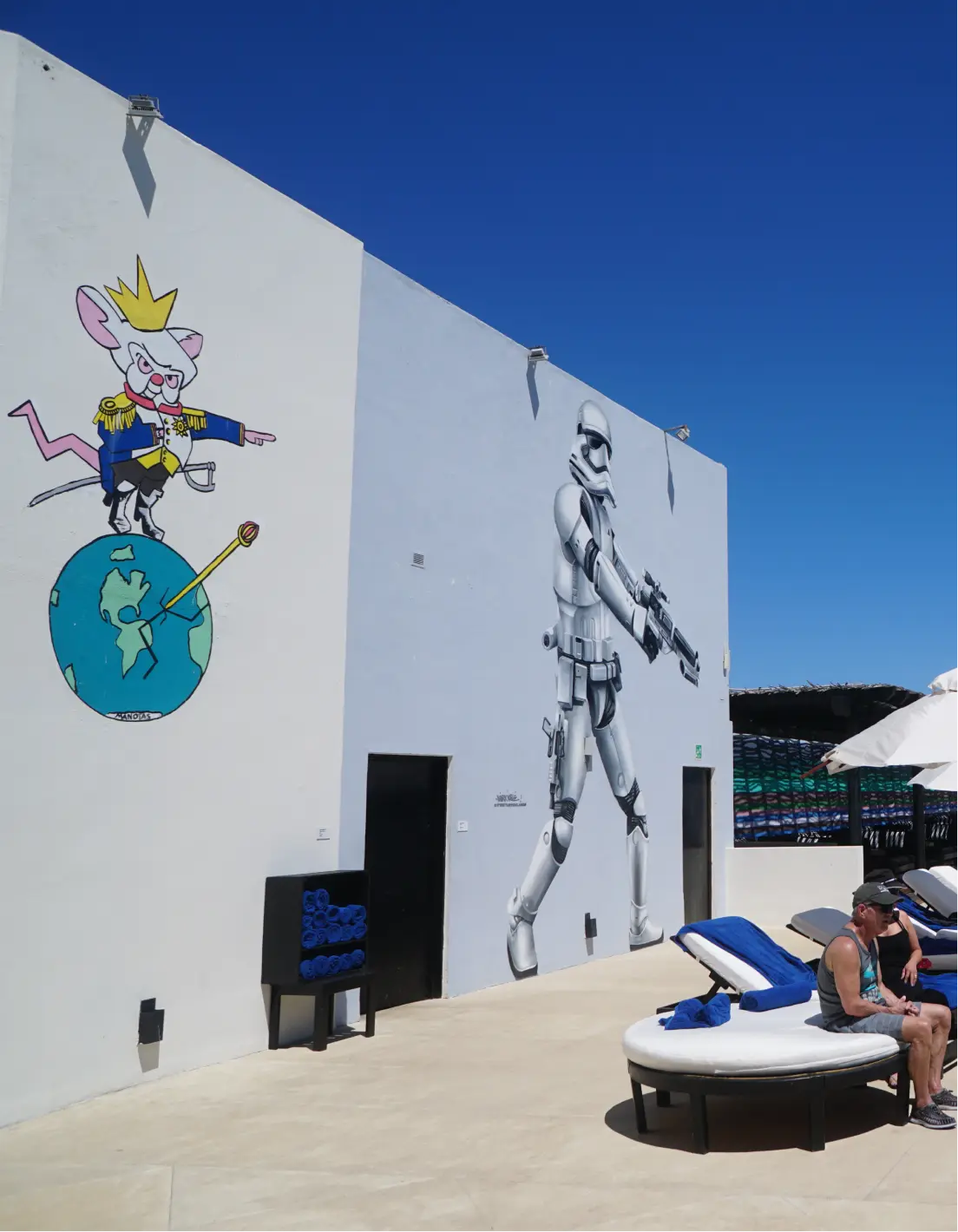 Murales exteriores que muestran a un rey ratón de dibujos animados de pie sobre un globo terráqueo y a un gran soldado imperial de Star Wars apuntando con un bláster, con tumbonas y gente relajándose cerca bajo un cielo azul claro.