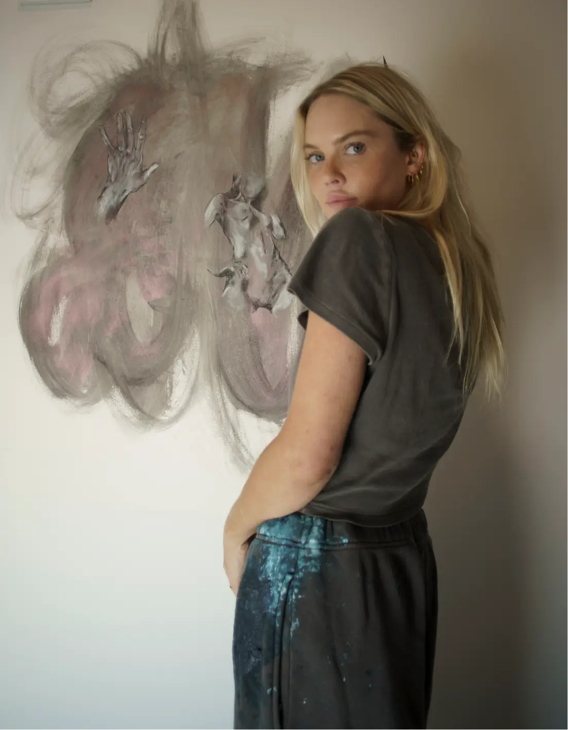 Mujer rubia de ojos claros con una camiseta oscura y pantalones salpicados de pintura, de pie frente a una pintura mural abstracta con bocetos de manos y torsos.