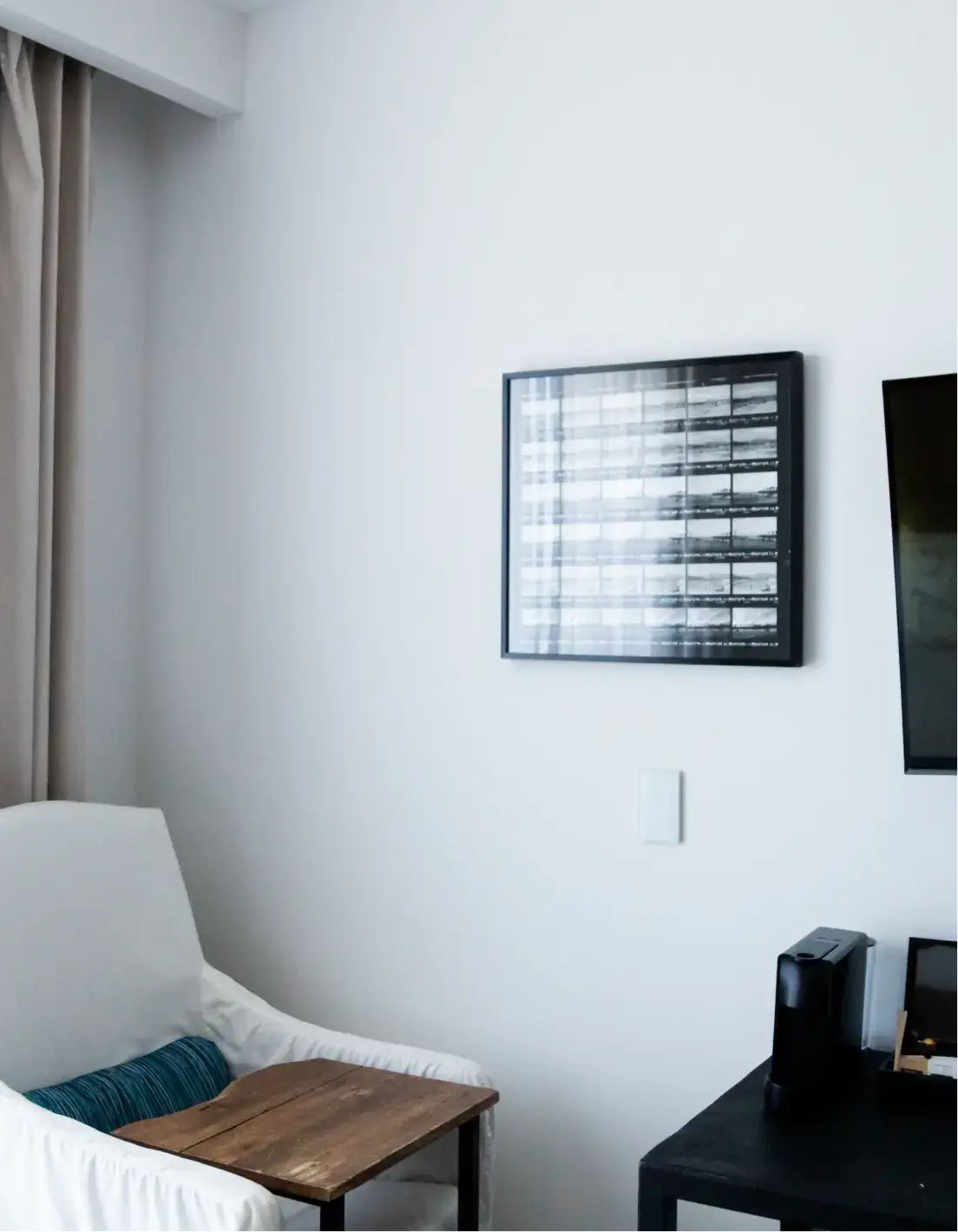 Sillón blanco con un cojín azul y una mesita auxiliar de madera junto a una pared con cuadros enmarcados y una cafetera negra sobre una mesa negra.
