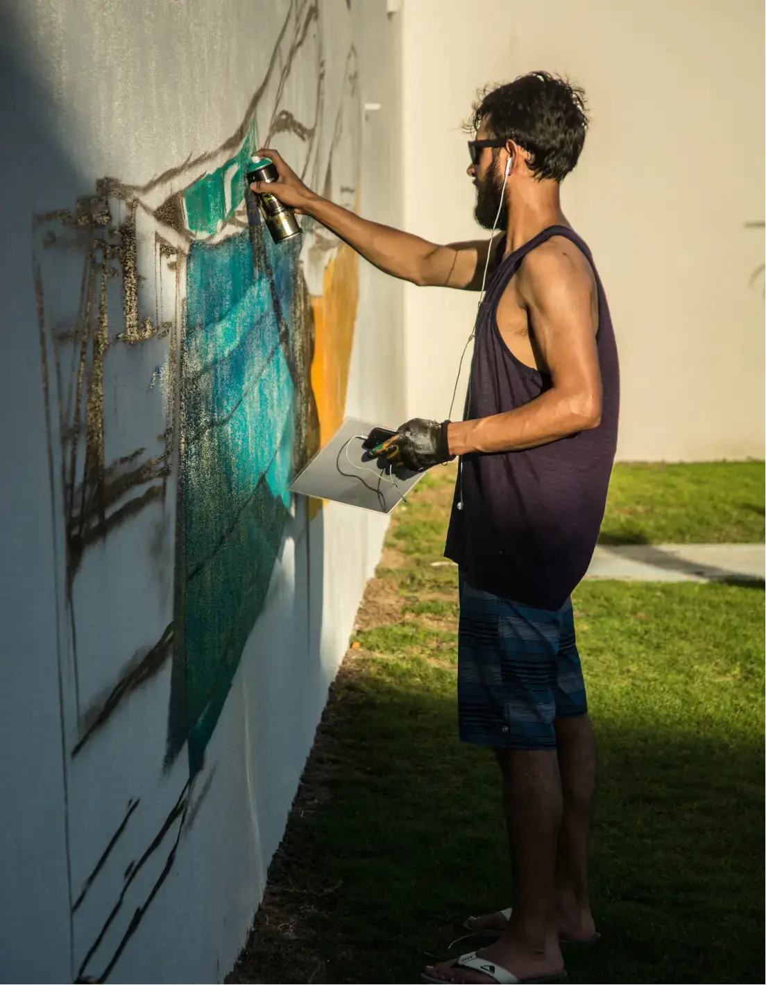 Hombre con gafas de sol y guantes pintando con spray un colorido mural en una pared blanca al aire libre bajo la luz del sol.