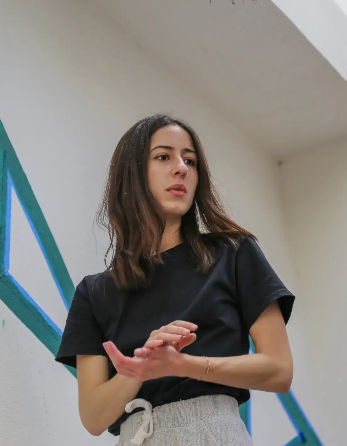 Mujer joven con cabello largo castaño, camiseta negra y pantalones gris claro, de pie en interiores cerca de una pared con rayas diagonales azules y verdes.