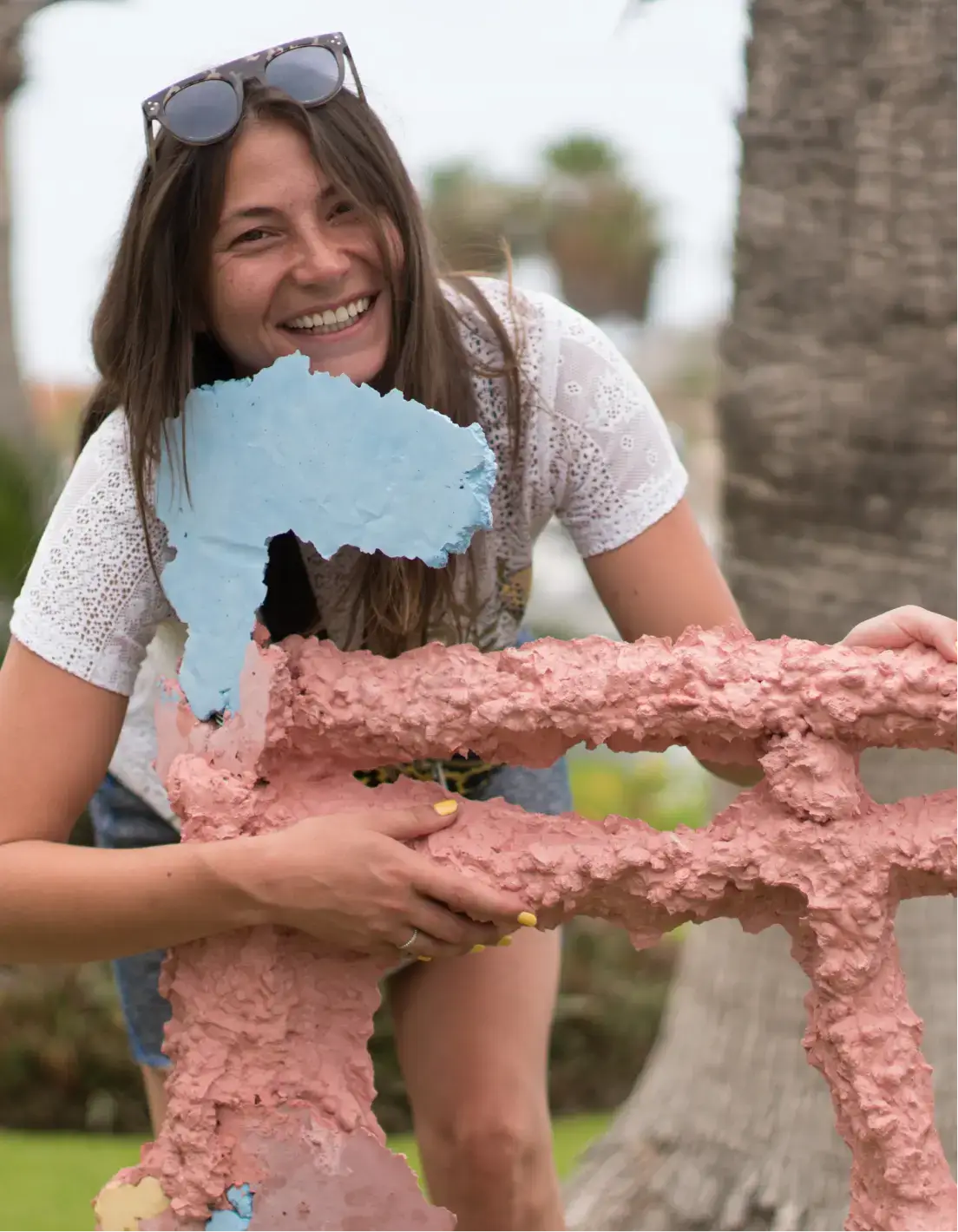 Mujer sonriente con gafas de sol en la cabeza sosteniendo una escultura exterior texturizada y colorida.