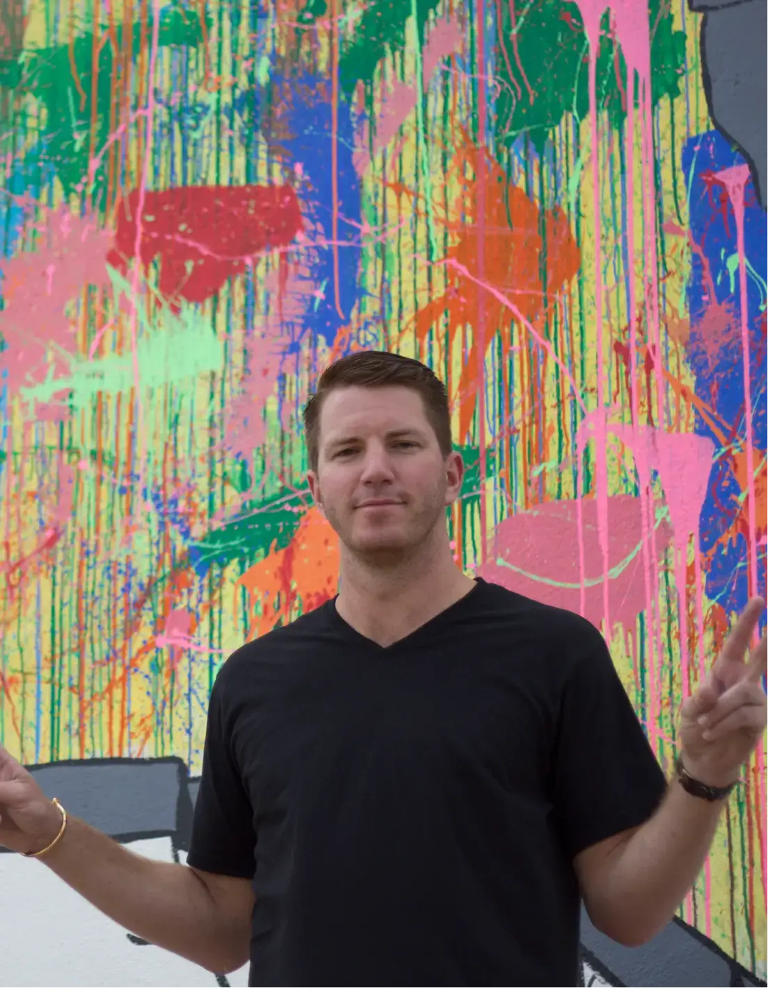 Hombre con camiseta negra posando con signos de paz delante de una pared abstracta salpicada de pintura de colores vivos.