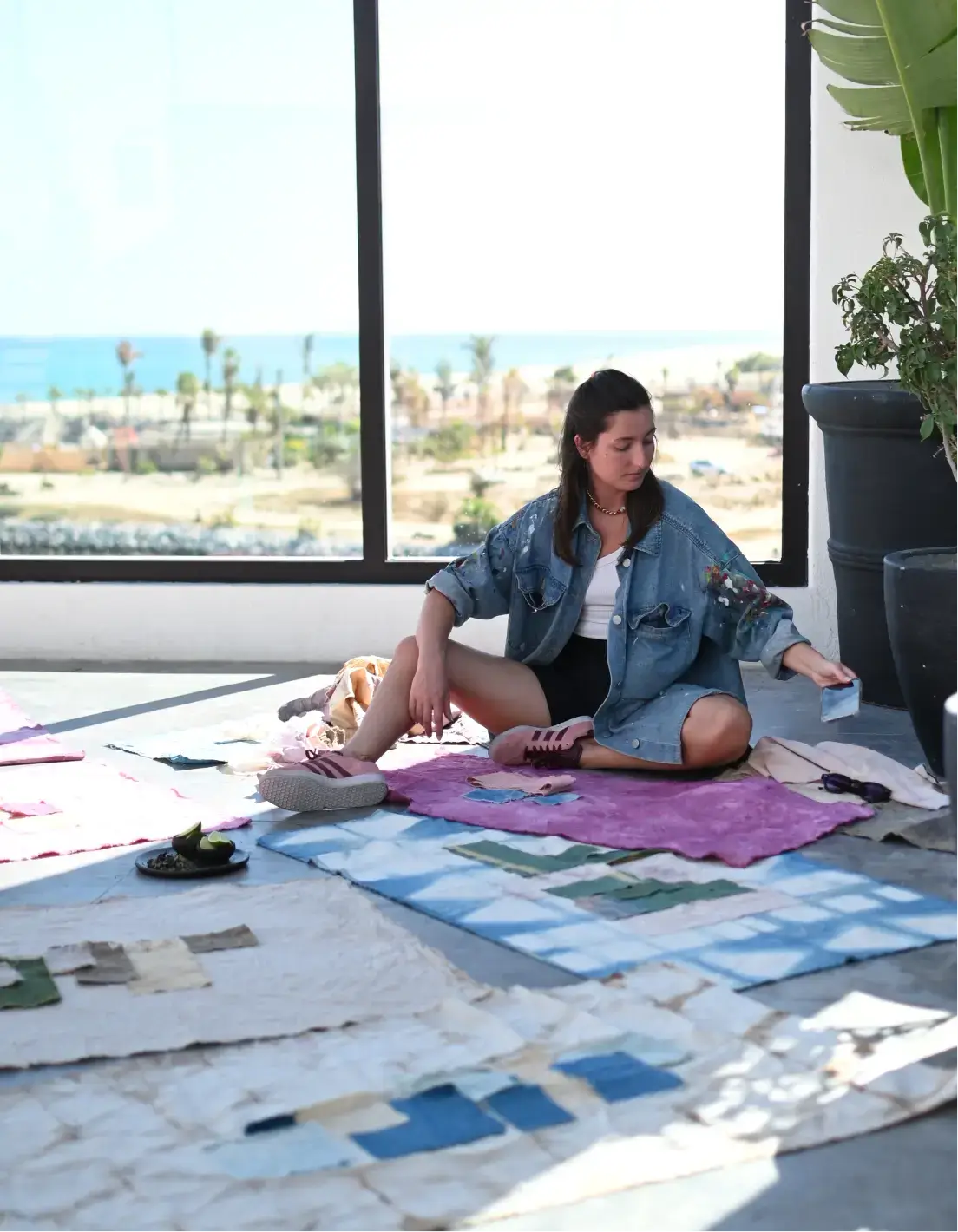 Mujer sentada en el suelo con trozos de tela de colores esparcidos, cerca de una gran ventana con vistas al mar.