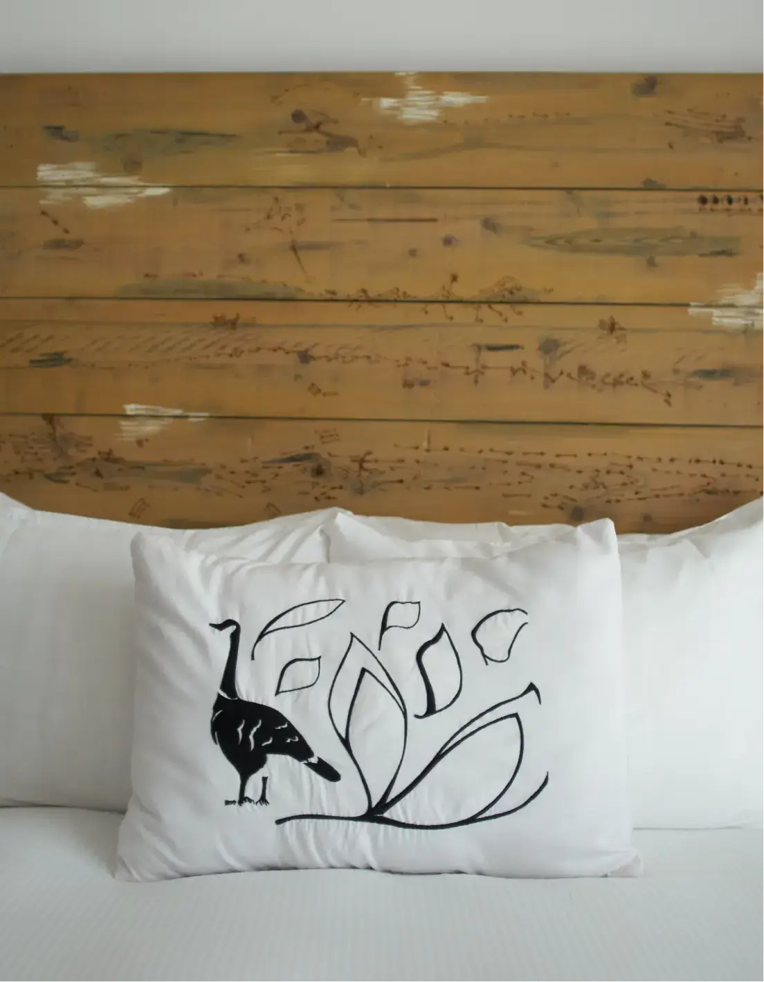 Almohada blanca sobre la cama con diseño de pájaro negro y hojas, cabecero de madera al fondo.