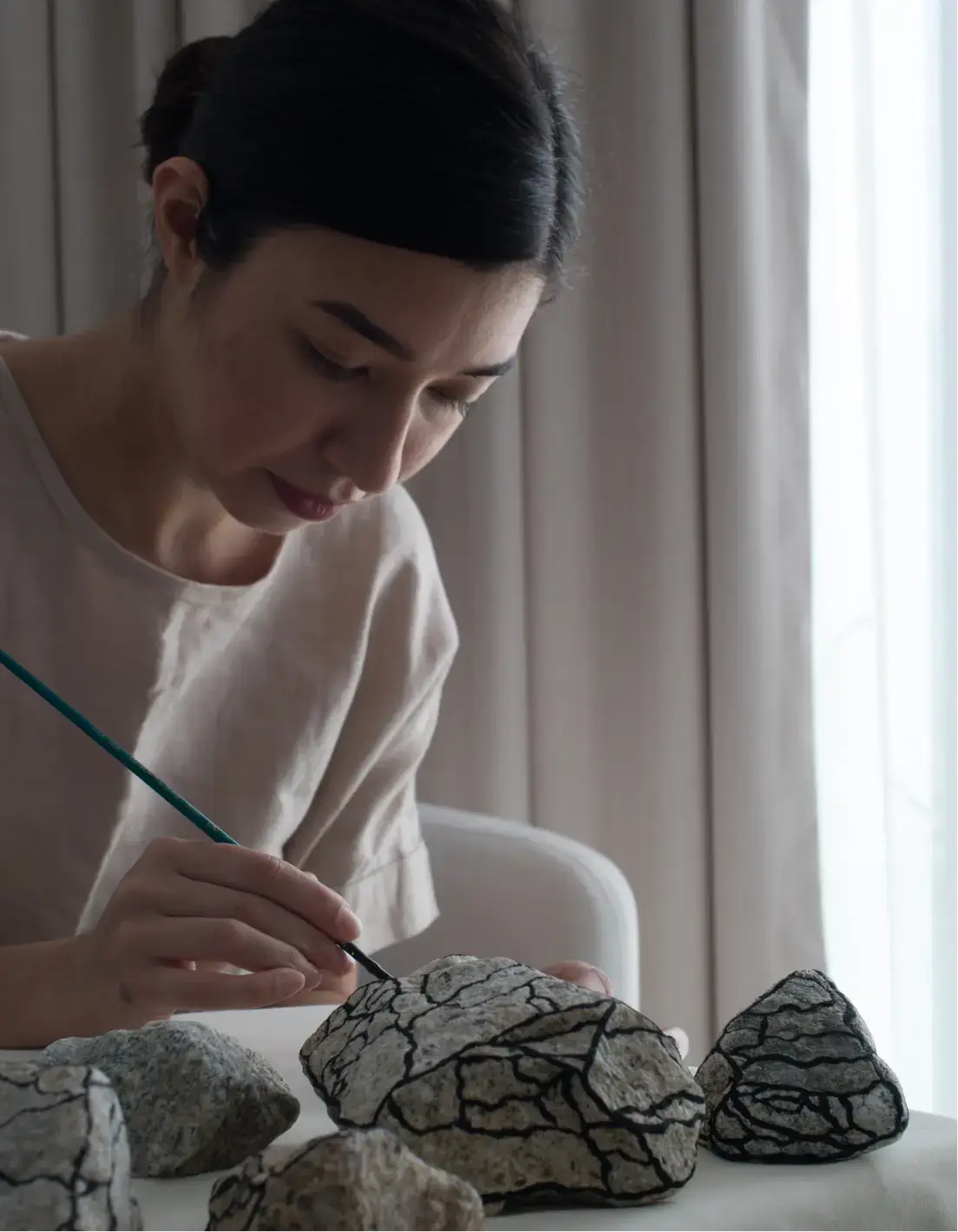 Mujer pintando líneas negras sobre una gran roca con un pincel fino en una mesa con otras rocas.
