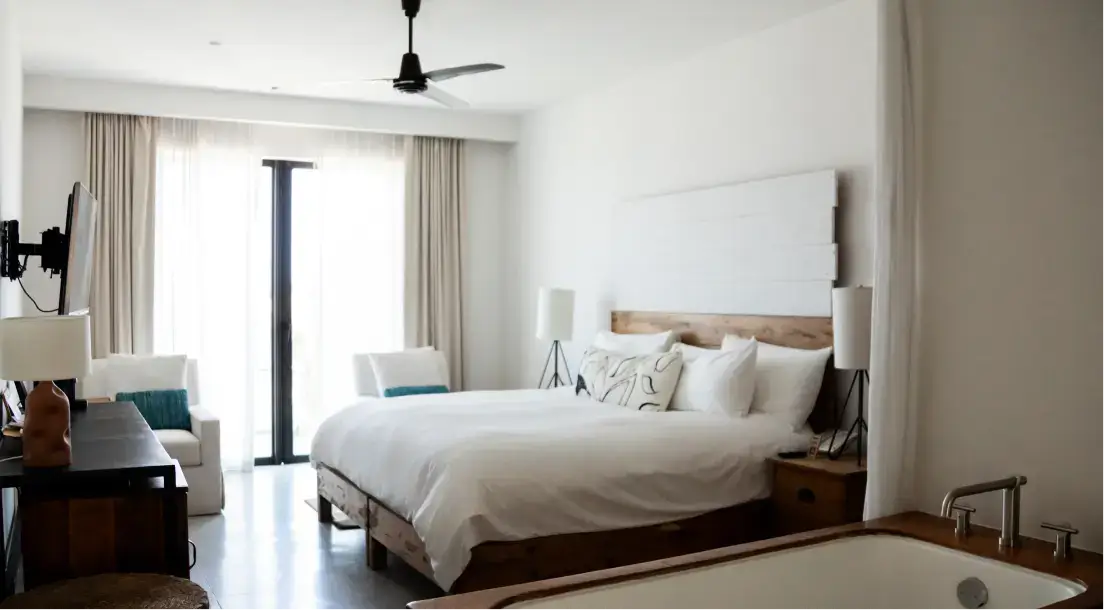 Bright bedroom with a white bed, two armchairs, wooden furniture, ceiling fan, and a bathtub in the foreground.