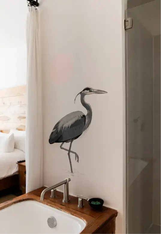 Lavabo de baño con encimera de madera y pintura mural de una garza junto a una ducha y una cortina blanca en un dormitorio.