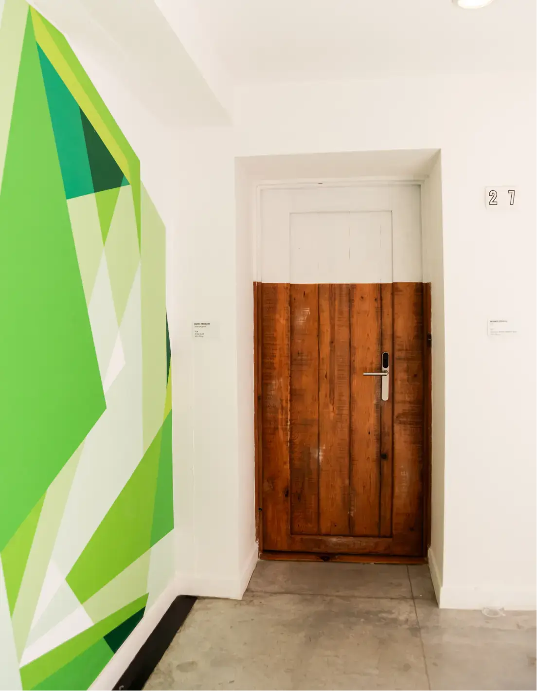 Interior hallway with a wooden door half painted white at the top, green geometric wall art on the left, and the number 27 on the right wall.
