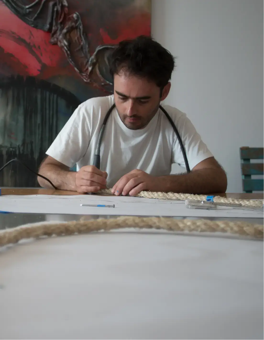 Man in white t-shirt using a tool to work on braided rope at a table indoors.