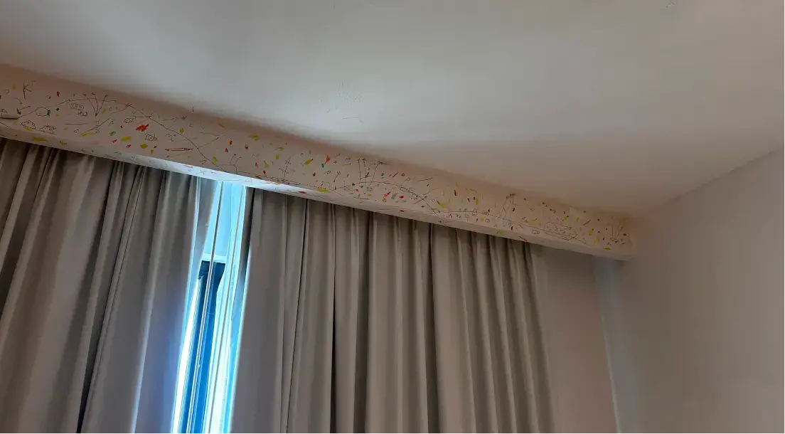 Light beige curtain covering a window with a decorative valance featuring colorful abstract line drawings and small shapes.