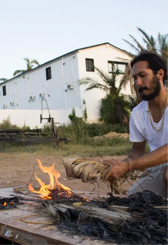 Man with a beard and long hair lighting a fire outdoors next to burnt and burning branches and leaves on a table.