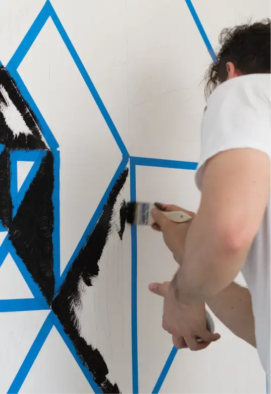 Person painting black geometric shapes within blue tape outlines on a white wall.