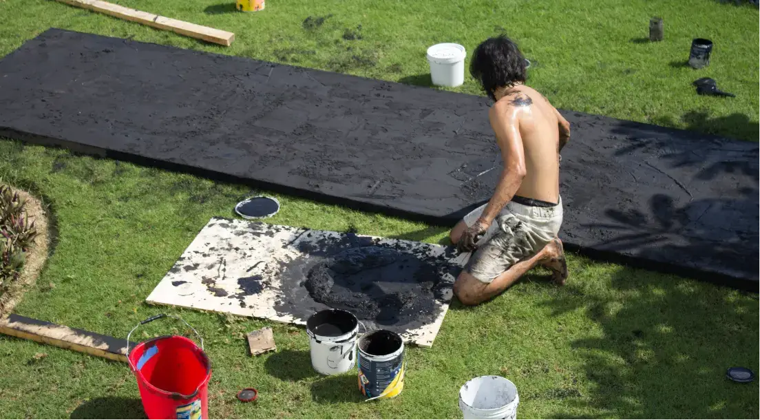 Shirtless man kneeling on grass mixing black paint or tar on a board surrounded by paint buckets.