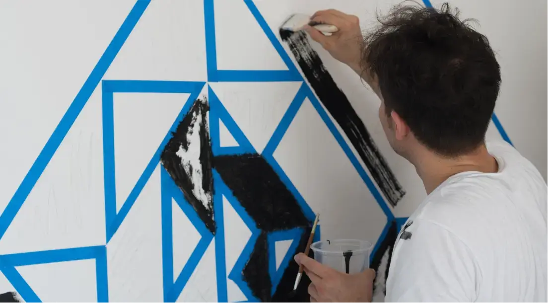 Man painting black shapes inside blue triangle outlines on a white wall.
