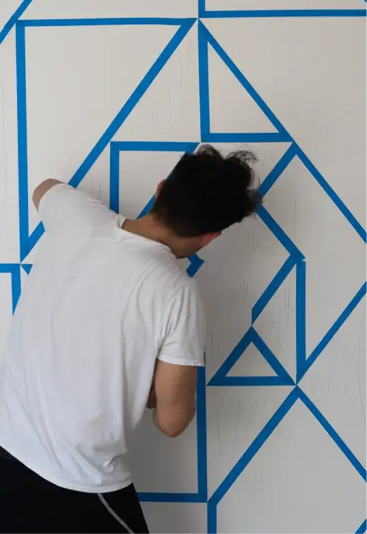 Person in a white t-shirt applying blue painter's tape in geometric shapes on a white wall.
