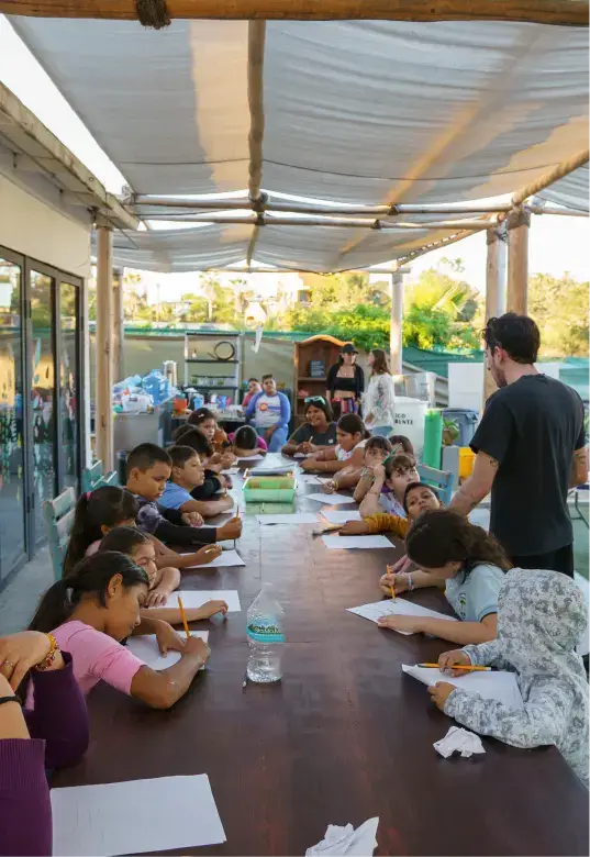 A group of children seated at a long outdoor table under a canopy, engaged in writing or drawing, while an adult stands nearby assisting.